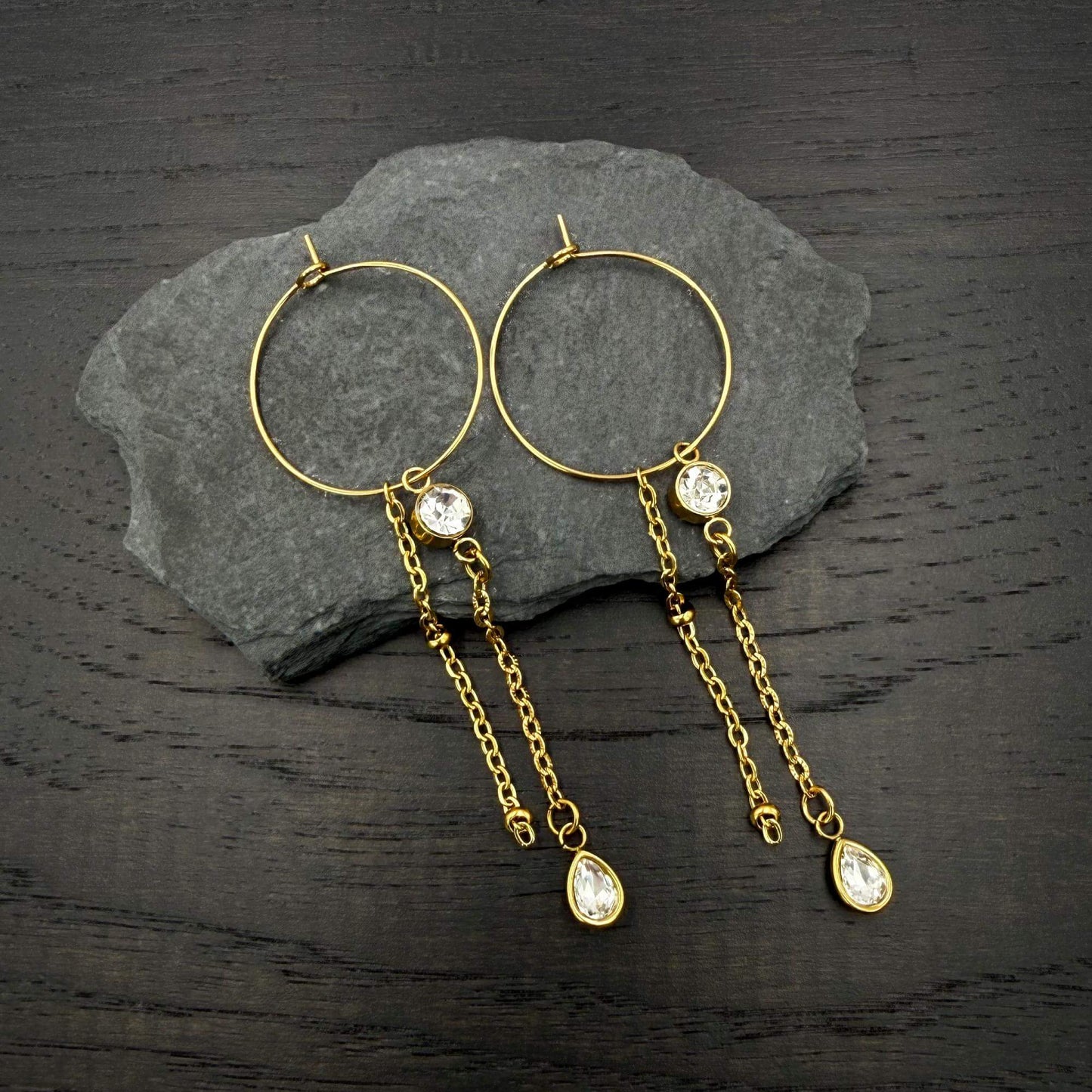 A pair of gold hoop earrings with dangling chain elements, clear faceted gemstones, and teardrop-shaped gold pendants, displayed on a dark gray slate slab over a dark wood surface.