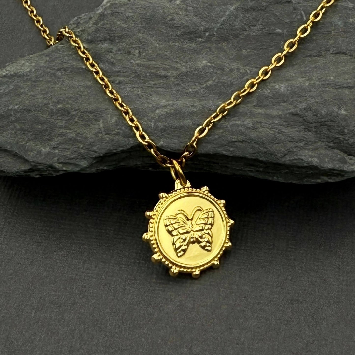 A gold pendant necklace featuring an embossed butterfly design inside a circular, beaded frame, displayed on a dark slate surface with a larger dark gray rock in the background.