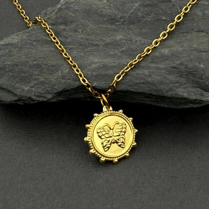A gold pendant necklace featuring an embossed butterfly design inside a circular, beaded frame, displayed on a dark slate surface with a larger dark gray rock in the background.