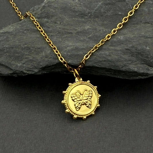 A gold pendant necklace featuring an embossed butterfly design inside a circular, beaded frame, displayed on a dark slate surface with a larger dark gray rock in the background.