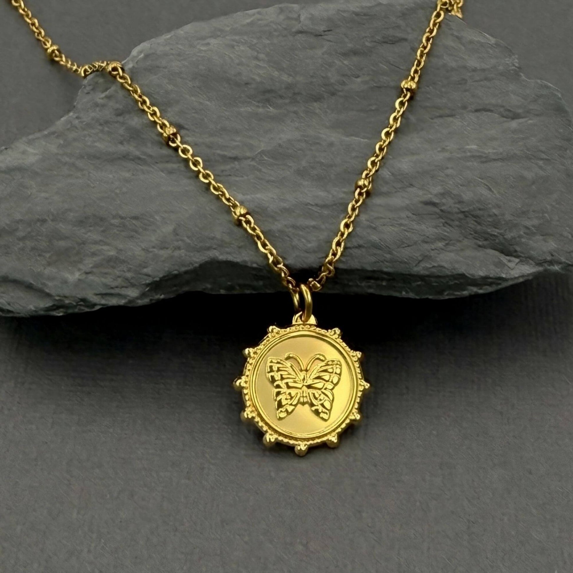 A gold pendant necklace featuring a circular medallion with an embossed butterfly design, displayed on a dark gray slate surface.
