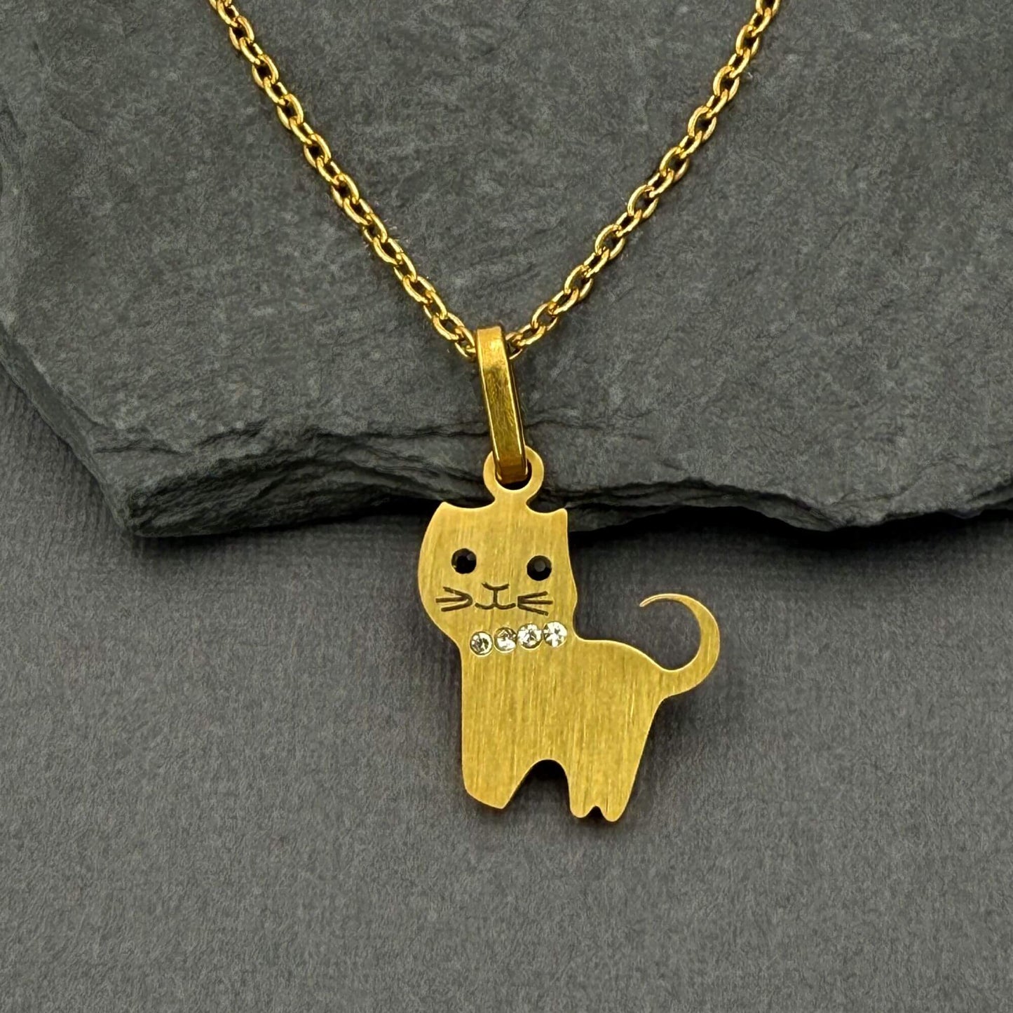A gold cat-shaped pendant necklace with small clear rhinestones on its neck, displayed on a dark gray slate surface. 