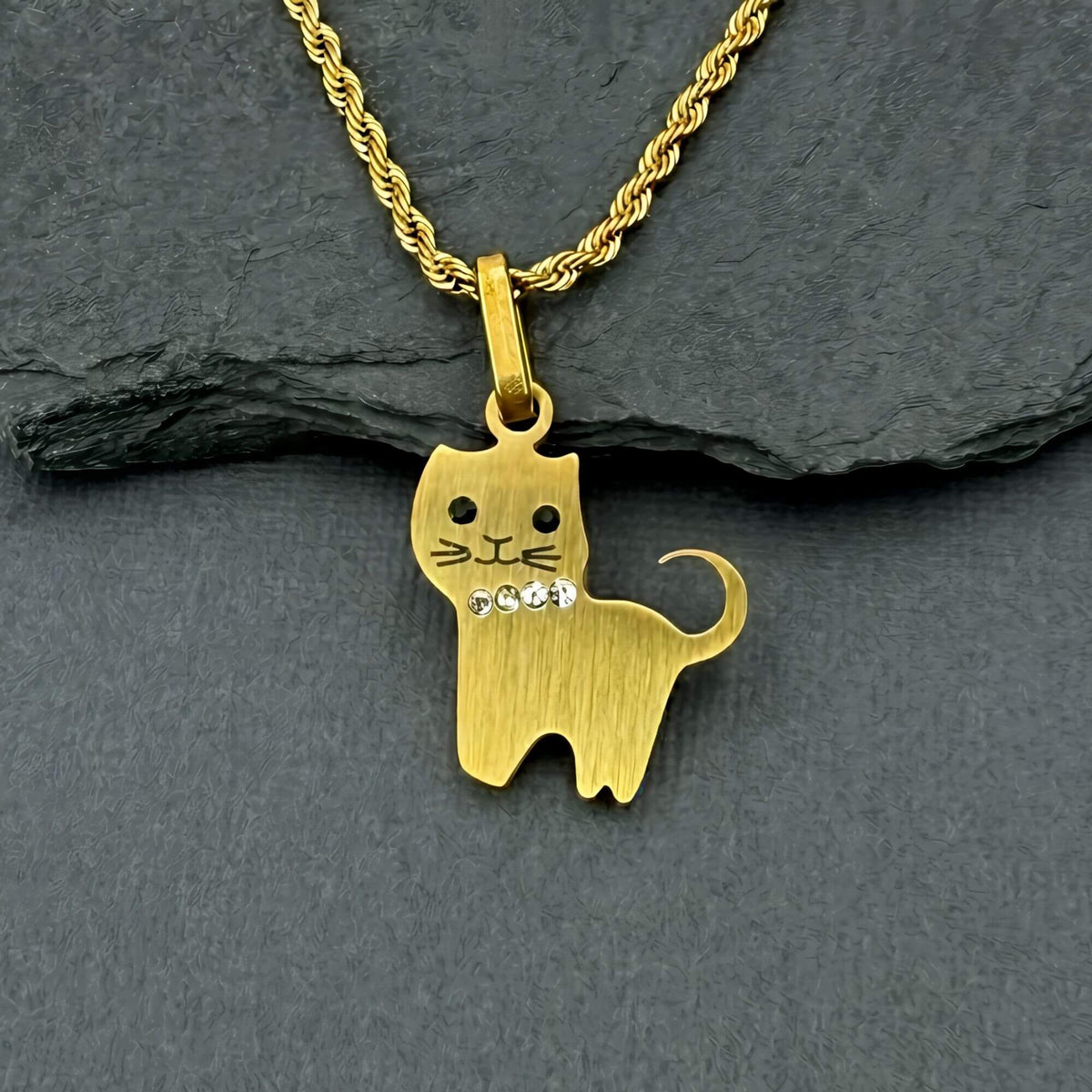A gold cat-shaped pendant necklace with a small rhinestone detail on its chest, resting on a dark gray slate surface.