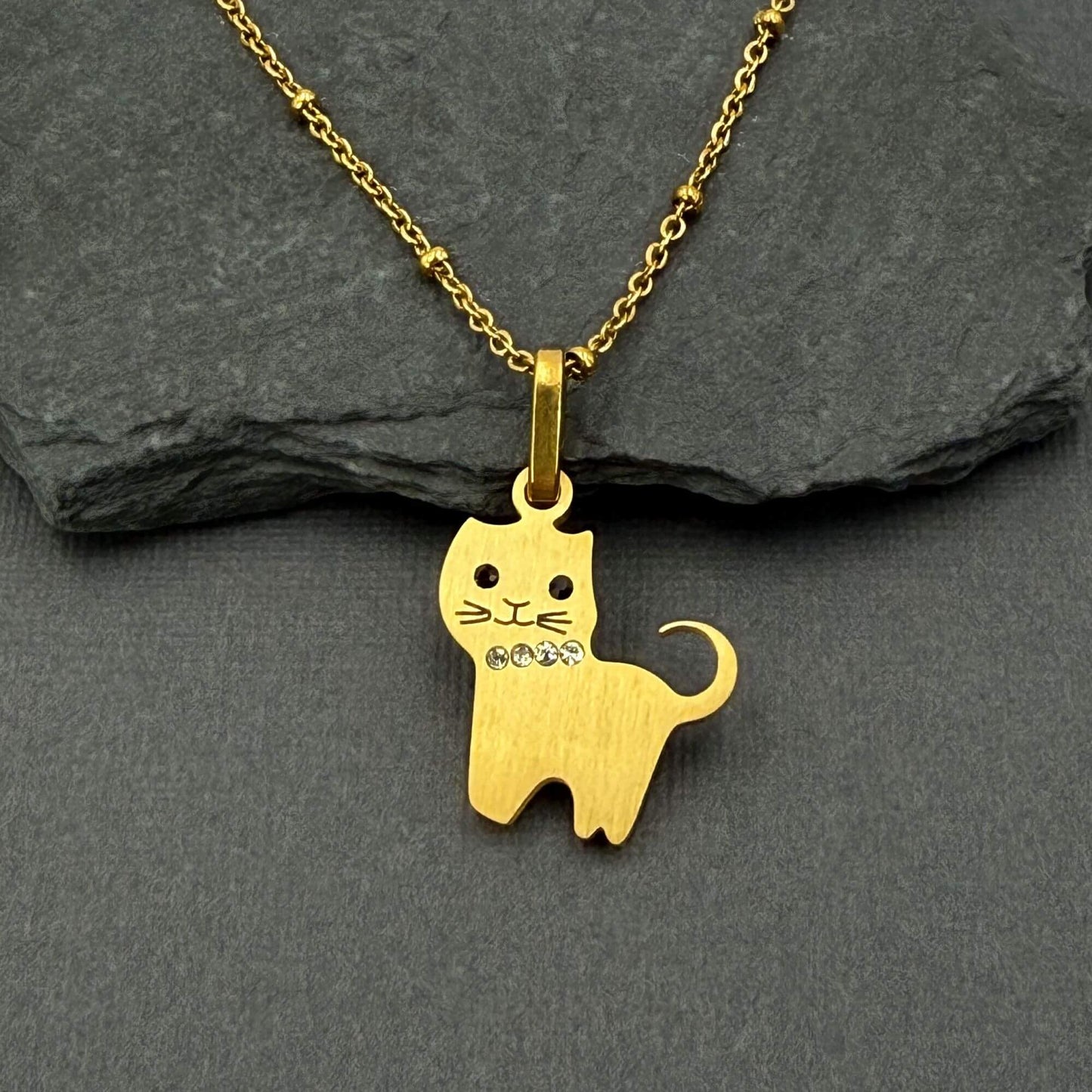 A gold cat-shaped pendant necklace with small rhinestone accents on its chest, displayed on a dark gray slate surface.