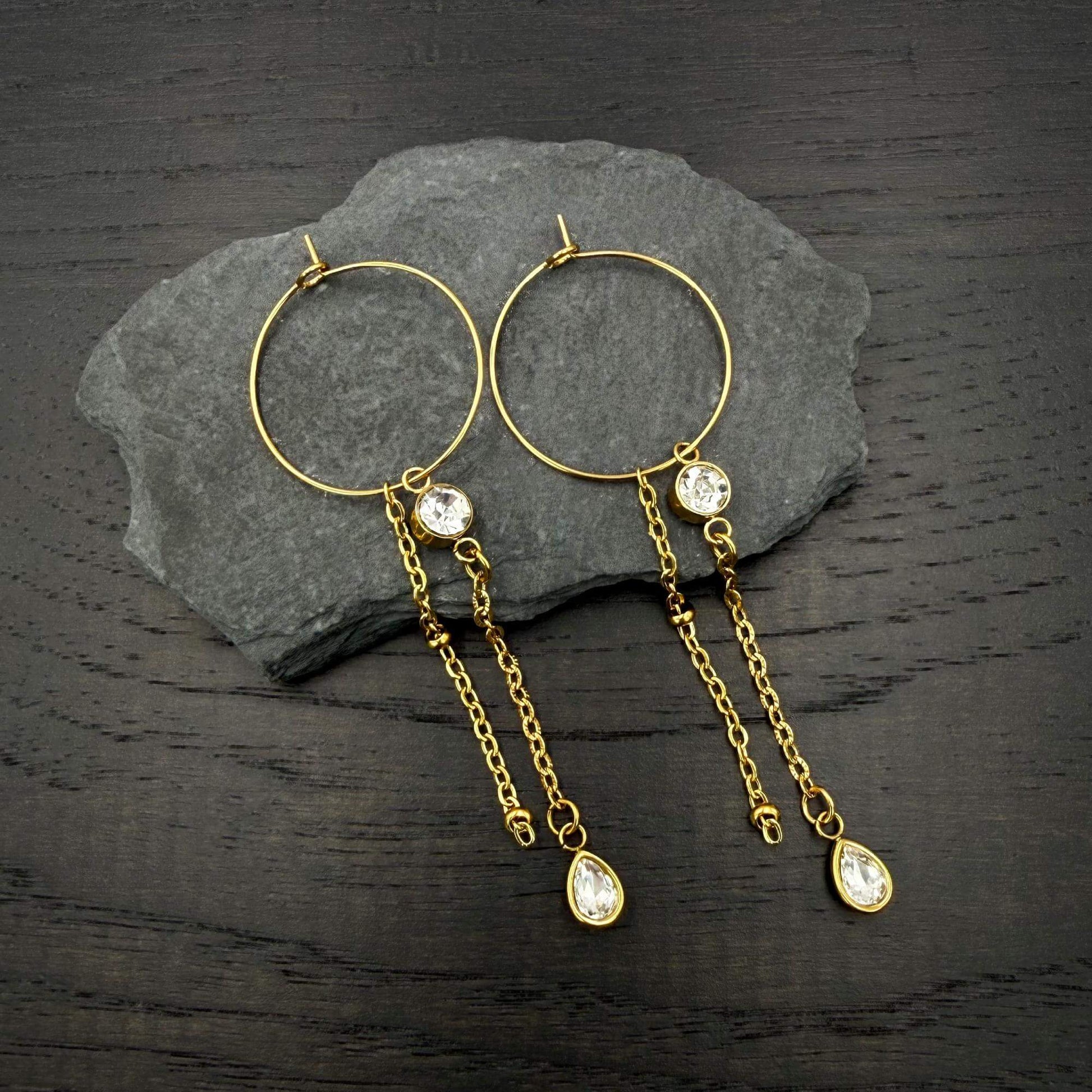 A pair of gold hoop earrings with dangling chain elements, clear faceted gemstones, and teardrop-shaped gold pendants, displayed on a dark gray slate slab over a dark wood surface.