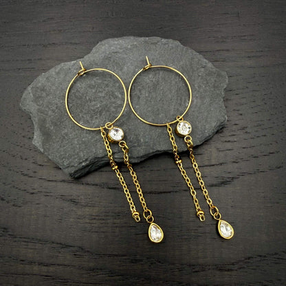 A pair of gold hoop earrings with dangling chain elements, clear faceted gemstones, and teardrop-shaped gold pendants, displayed on a dark gray slate slab over a dark wood surface.