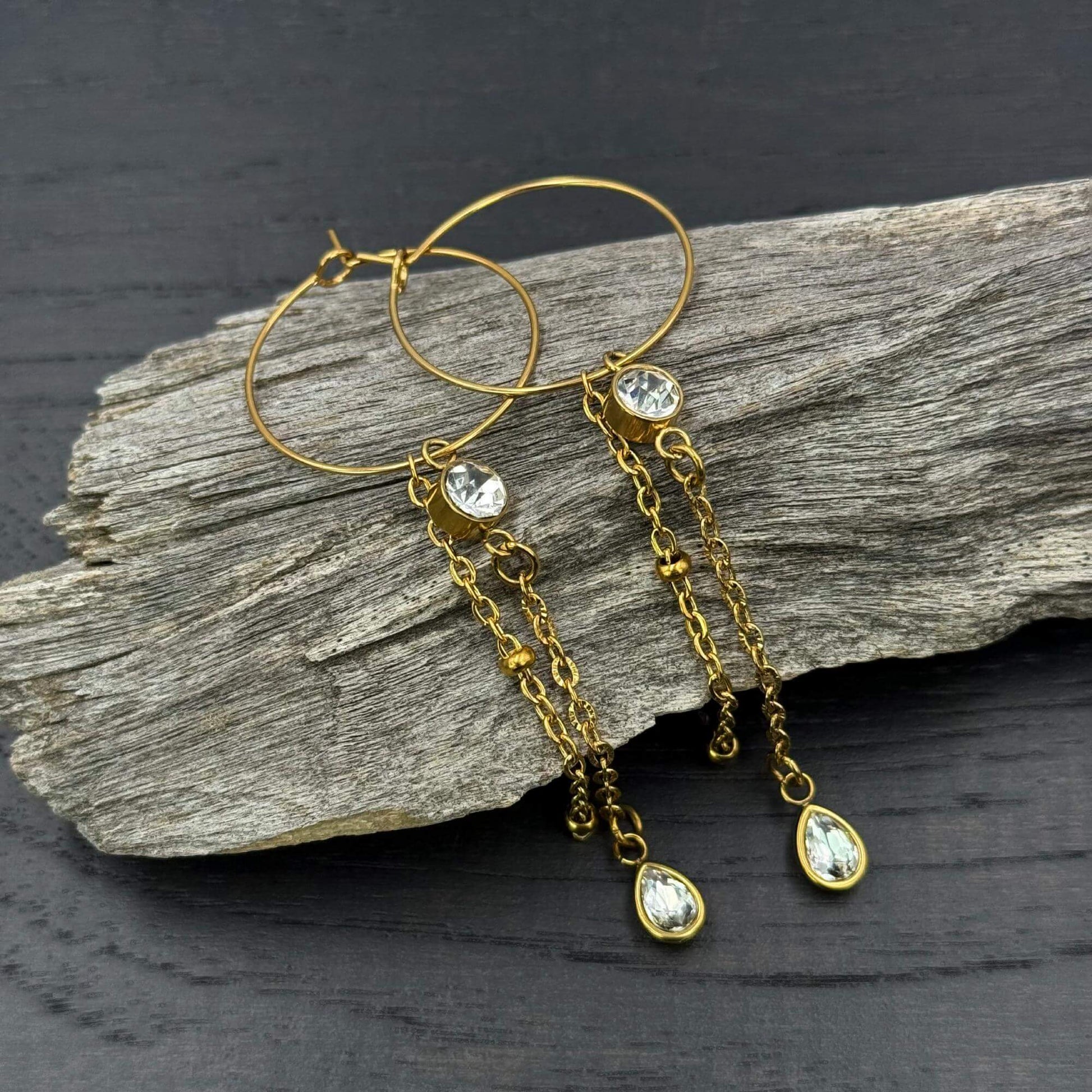 A pair of gold hoop chandelier chain dangle earrings with clear teardrop gemstones and dangling chain accents, displayed on a piece of weathered driftwood over a dark textured surface.