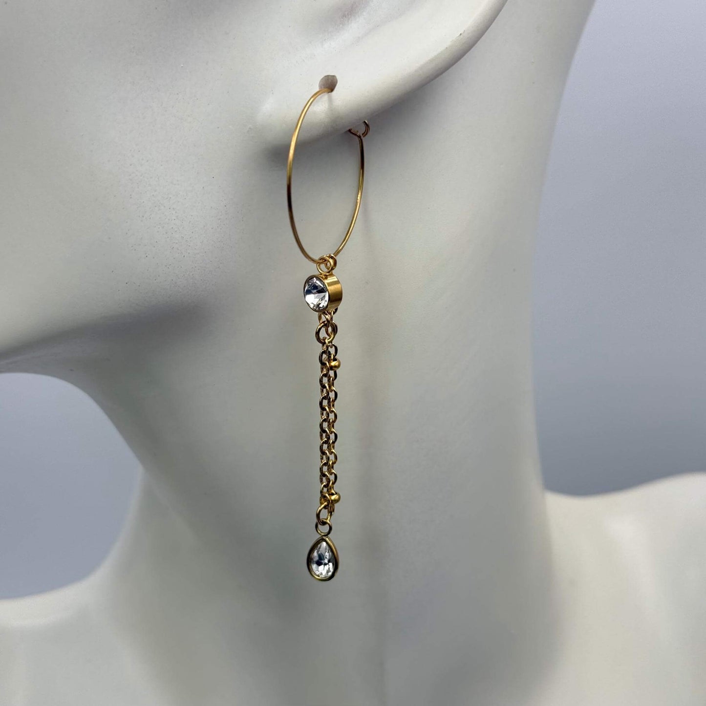 A gold hoop chandelier dangle earring with a clear teardrop gemstone, displayed on a white mannequin head against a soft gray background.