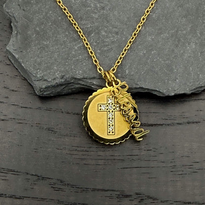 A gold pendant necklace featuring a round scalloped disc with a cross and small clear stones, alongside a dangling script charm that reads 'Jesus is the Lord,' displayed on a dark wood surface with a gray slate prop.