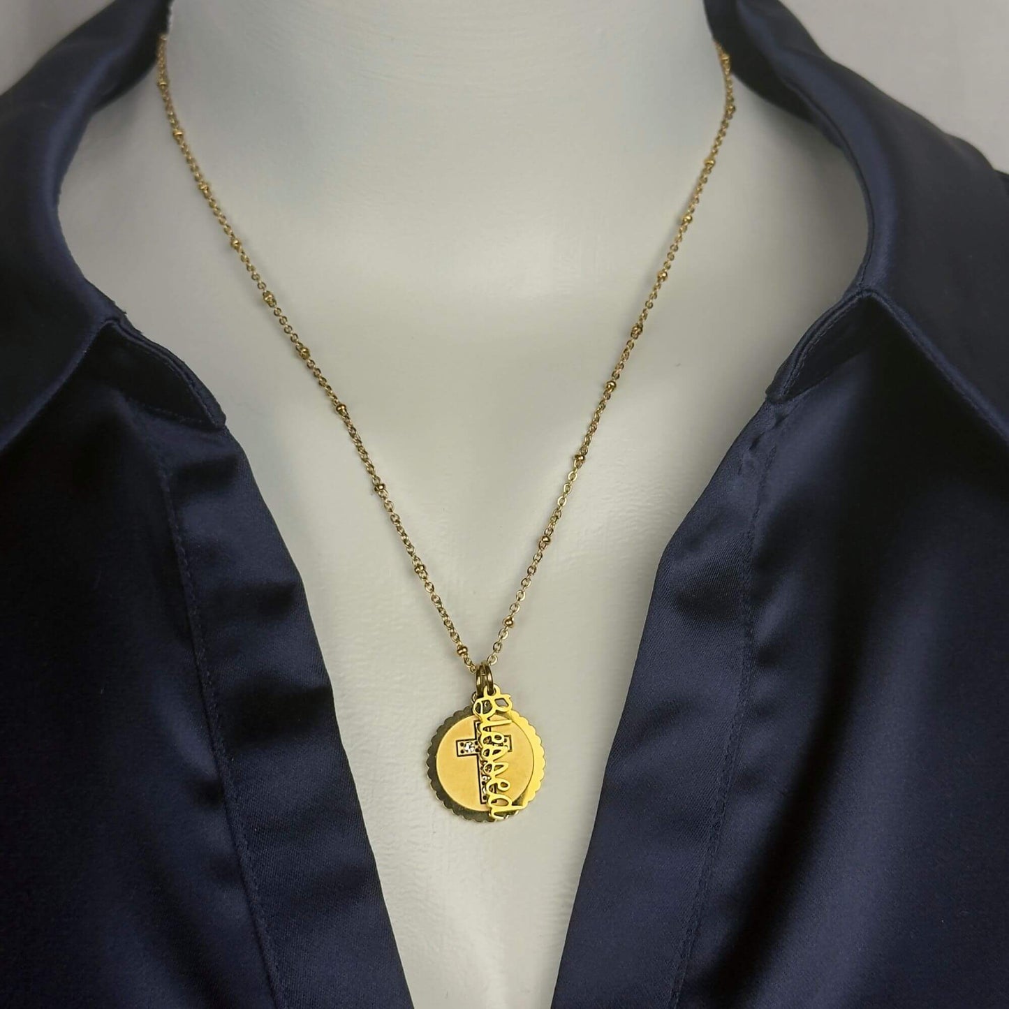 A gold pendant necklace displayed on a white mannequin torso wearing a navy satin shirt. The necklace features a delicate gold chain and a round gold pendant with a textured, engraved design.
