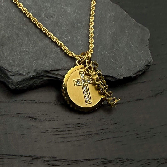 A gold necklace with a round pendant featuring a cross embellished with small clear stones, displayed on a dark textured surface next to a dark slate stone.