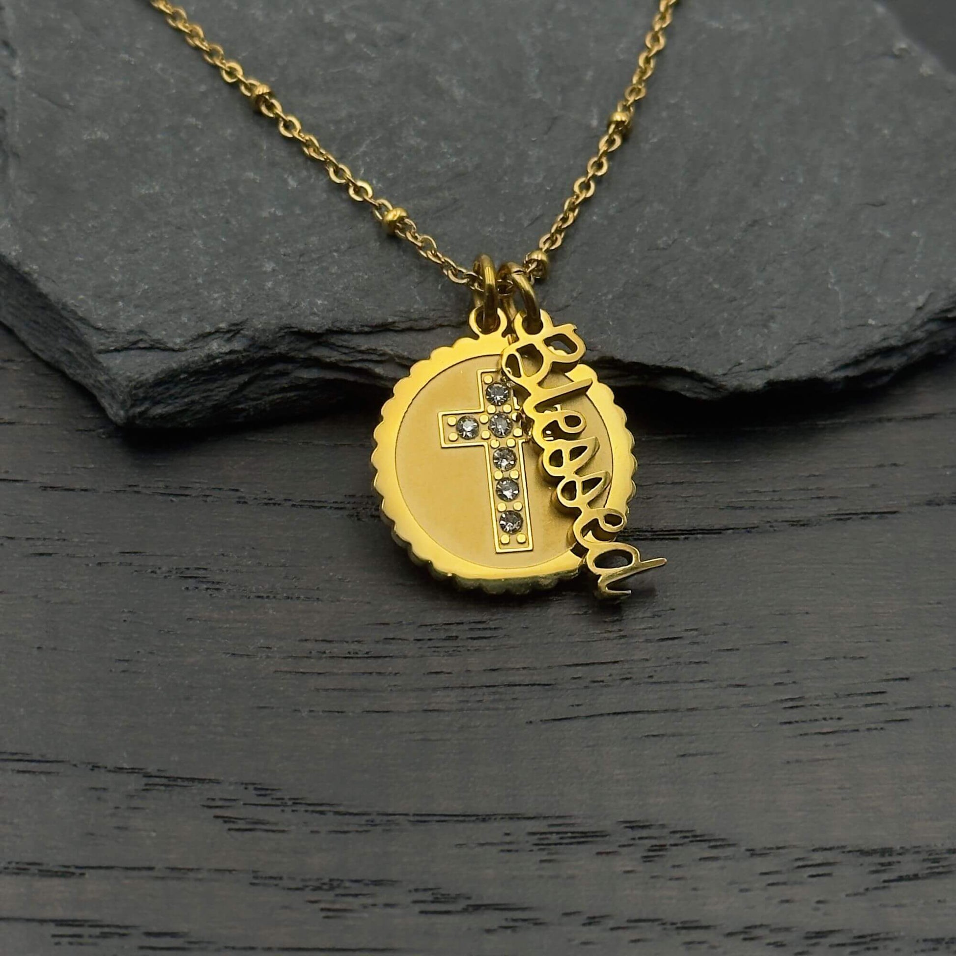 A gold necklace with a round pendant featuring a cross embellished with small clear stones, alongside a dangling script charm spelling 'Jesus', displayed on a dark textured surface.