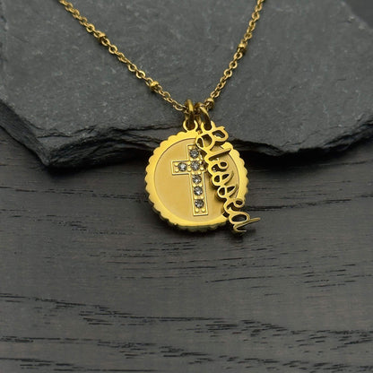 A gold necklace with a round pendant featuring a cross embellished with small clear stones, alongside a dangling script charm spelling 'Jesus', displayed on a dark textured surface.