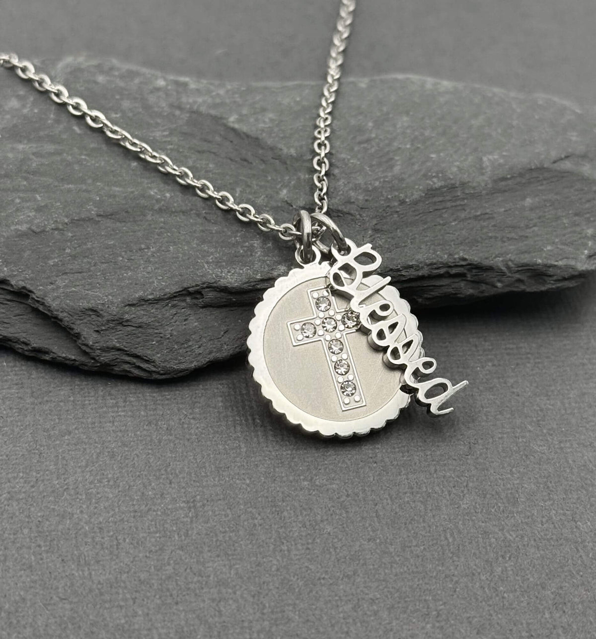 A silver stainless steel necklace with a round scalloped pendant, a small cross with clear rhinestones, and a dangling script charm spelling 'Blessed', displayed on a dark gray slate surface.