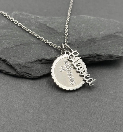 A silver stainless steel necklace with a round scalloped pendant, a small cross with clear rhinestones, and a dangling script charm spelling 'Blessed', displayed on a dark gray slate surface.