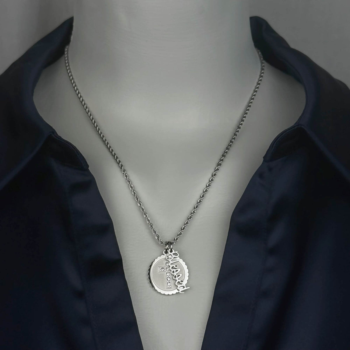 A stainless steel silver-toned chain necklace with a circular cross pendant engraved with the word 'Blessed', displayed on a mannequin torso wearing a dark navy satin shirt.