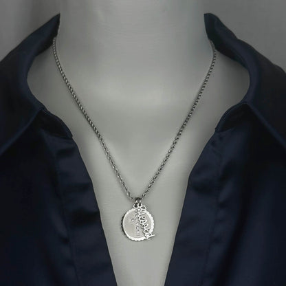 A stainless steel silver-toned chain necklace with a circular cross pendant engraved with the word 'Blessed', displayed on a mannequin torso wearing a dark navy satin shirt.