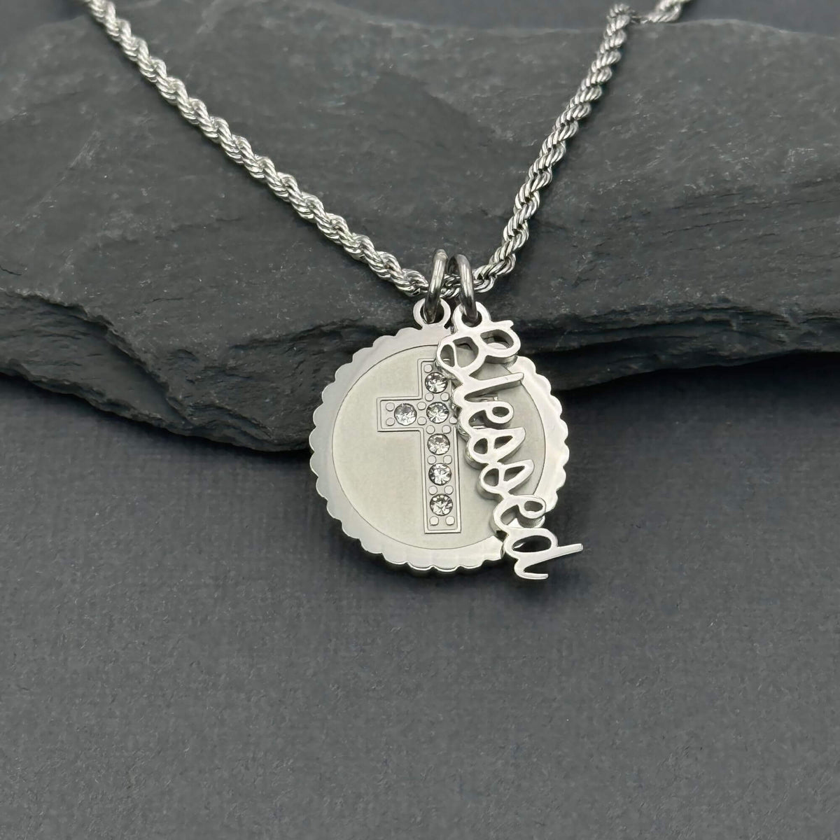 A silver stainless steel necklace with a rope chain and a round scalloped pendant featuring a cross with small clear crystal stones and a dangling cursive nameplate Blessed, displayed on a dark slate surface.