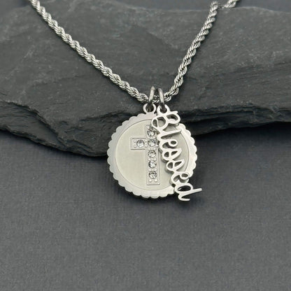 A silver stainless steel necklace with a rope chain and a round scalloped pendant featuring a cross with small clear crystal stones and a dangling cursive nameplate Blessed, displayed on a dark slate surface.