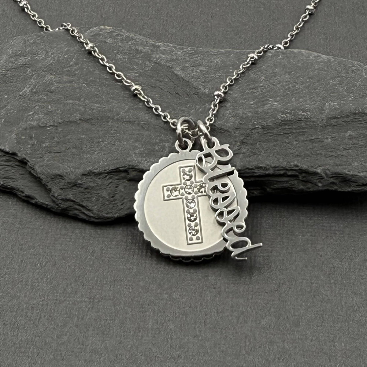 A silver stainless steel necklace with a round medallion pendant featuring a cross set with small clear stones, alongside a dangling script charm that reads 'Blessed', displayed on a dark gray slate surface.