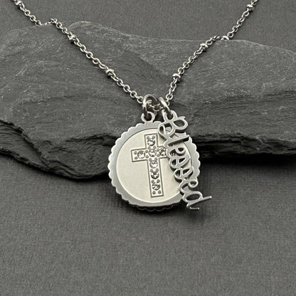 A silver stainless steel necklace with a round medallion pendant featuring a cross set with small clear stones, alongside a dangling script charm that reads 'Blessed', displayed on a dark gray slate surface.