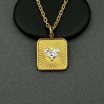 A gold square pendant necklace with a sunburst design and two clear heart-shaped gemstones in the center, displayed on a black circular stand against a dark gray background.