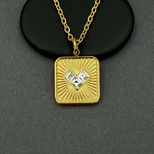 A gold square pendant necklace with a sunburst design and two clear heart-shaped gemstones in the center, displayed on a black circular stand against a dark gray background.