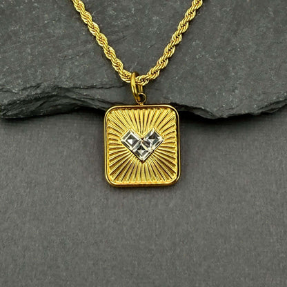 A gold square pendant necklace with a sunburst design and a central clear heart-shaped gemstone, resting on a dark gray slate surface.