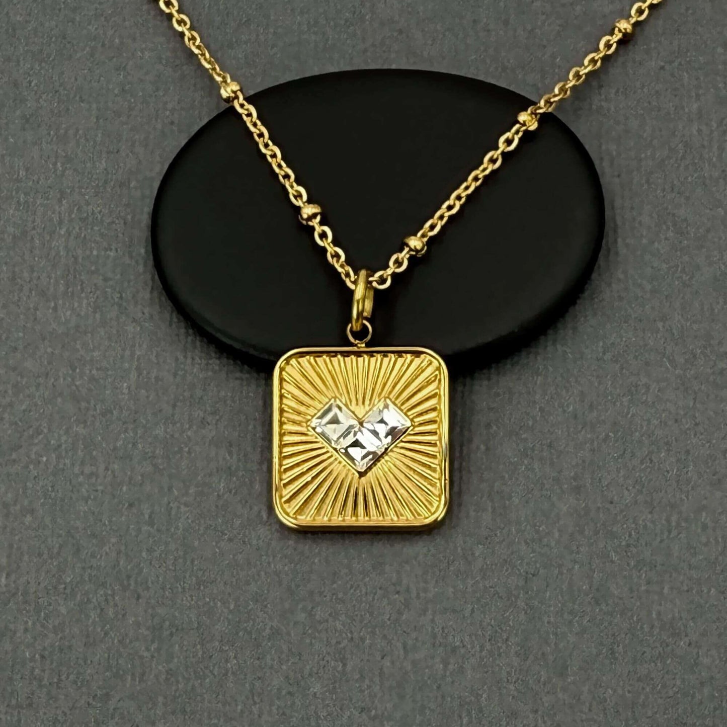 A gold square pendant necklace with a sunburst design and a central clear gemstone, displayed on a black oval stand against a dark gray background.
