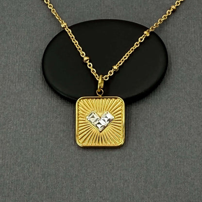 A gold square pendant necklace with a sunburst design and a central clear gemstone, displayed on a black oval stand against a dark gray background.