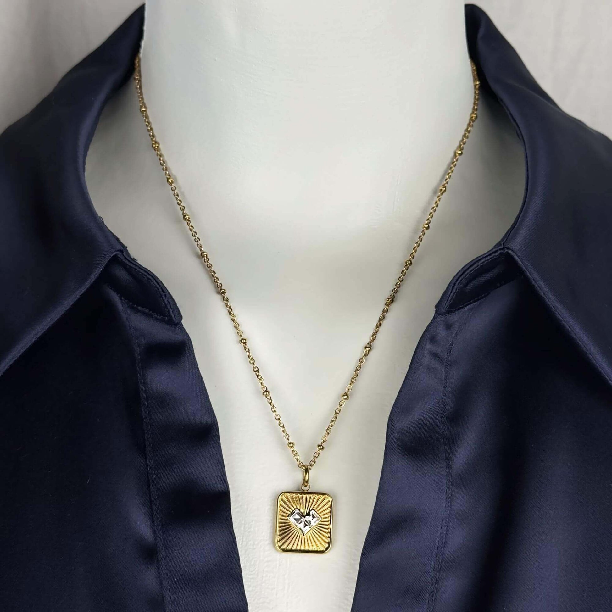 A gold square pendant necklace with a sunburst design and a small central heart, displayed on a white mannequin torso wearing a navy satin shirt.