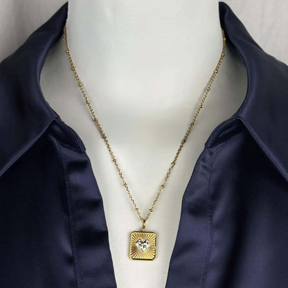 A gold square pendant necklace with a sunburst design and a small central heart, displayed on a white mannequin torso wearing a navy satin shirt.
