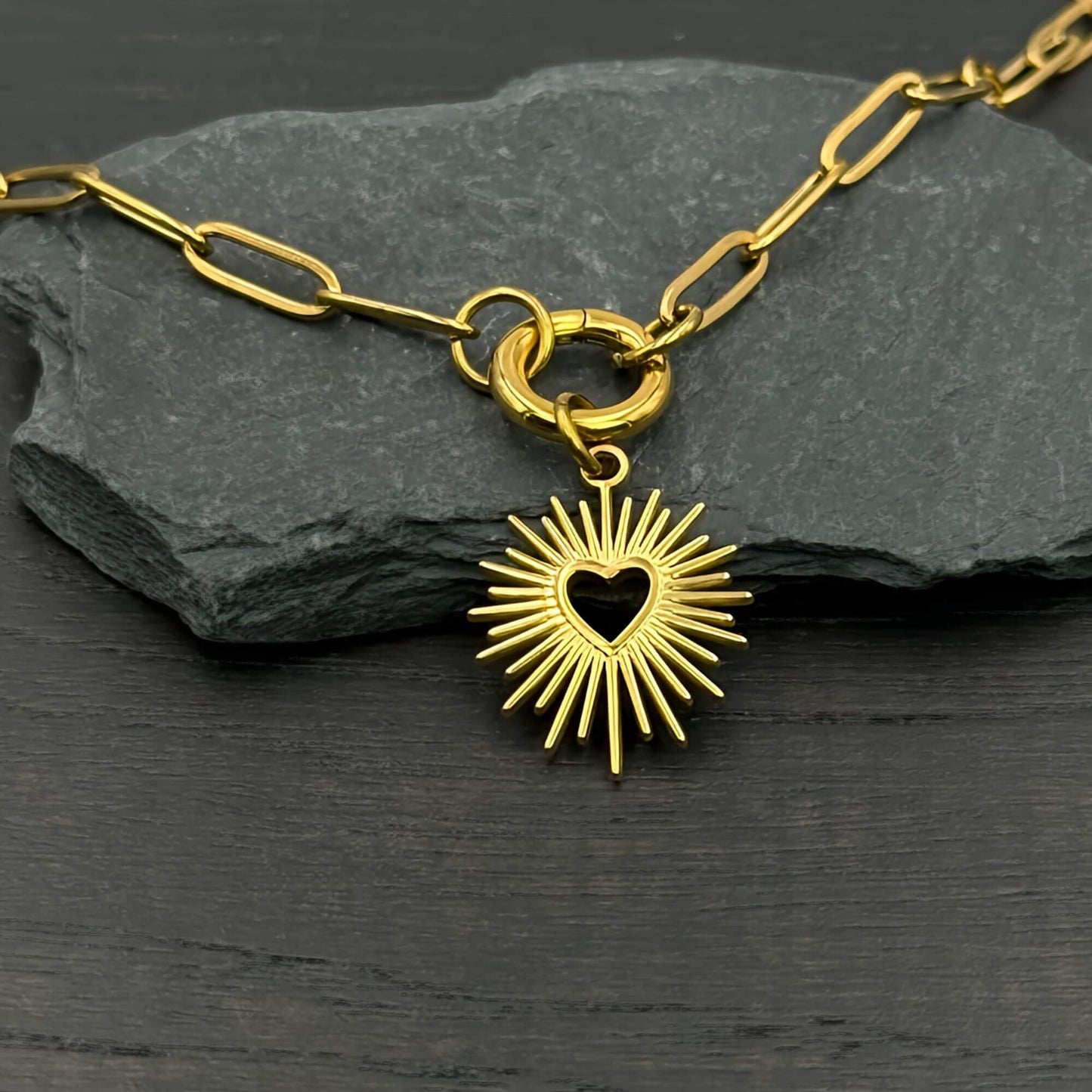 A gold-toned paperclip chain necklace with a heart-shaped sunburst pendant, displayed on a dark slate rock over a dark wooden surface.