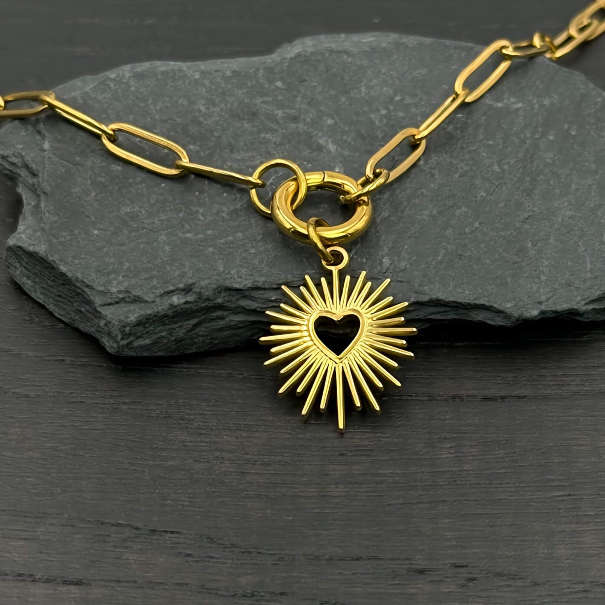 A gold-toned paperclip chain necklace with a heart-shaped sunburst pendant, displayed on a dark slate rock over a dark wooden surface.