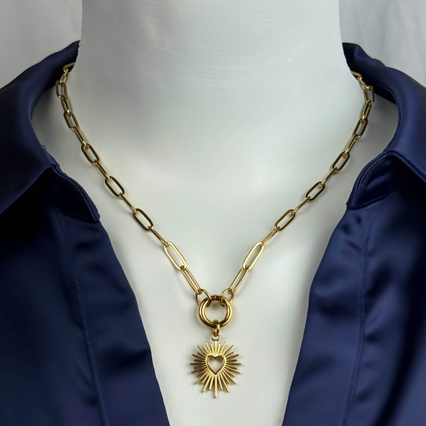 A gold-toned paperclip chain necklace with a central ring and a sunburst heart pendant, displayed on a white mannequin torso wearing a navy satin shirt.