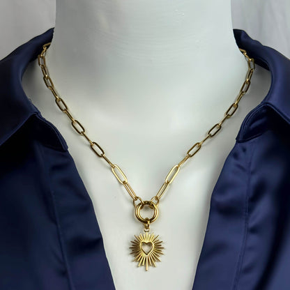 A gold-toned paperclip chain necklace with a central ring and a sunburst heart pendant, displayed on a white mannequin torso wearing a navy satin shirt.