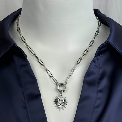 A silver paperclip chain necklace with a heart-shaped sun pendant, displayed on a white mannequin torso wearing a navy blue satin shirt.