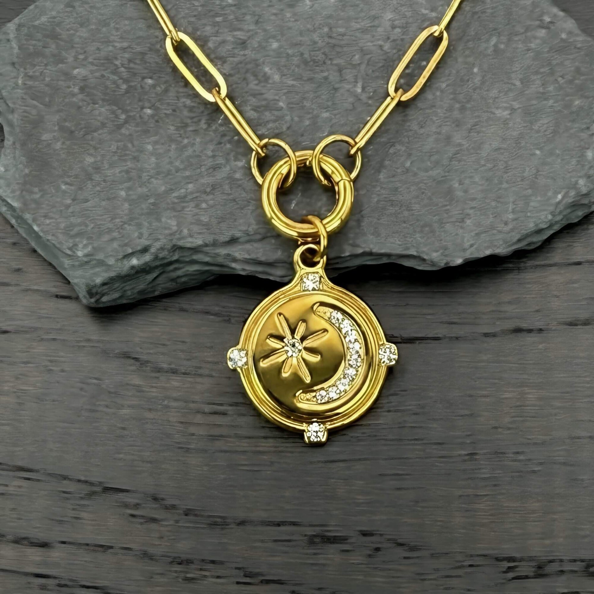 A gold-toned paperclip chain pendant necklace featuring a circular medallion with a crescent moon, star, and small clear stones, displayed on a dark gray slate slab over a dark wood surface.
