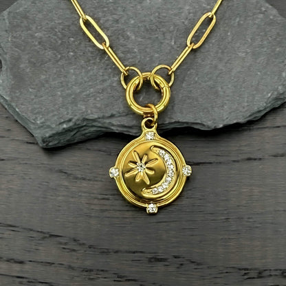 A gold-toned paperclip chain pendant necklace featuring a circular medallion with a crescent moon, star, and small clear stones, displayed on a dark gray slate slab over a dark wood surface.