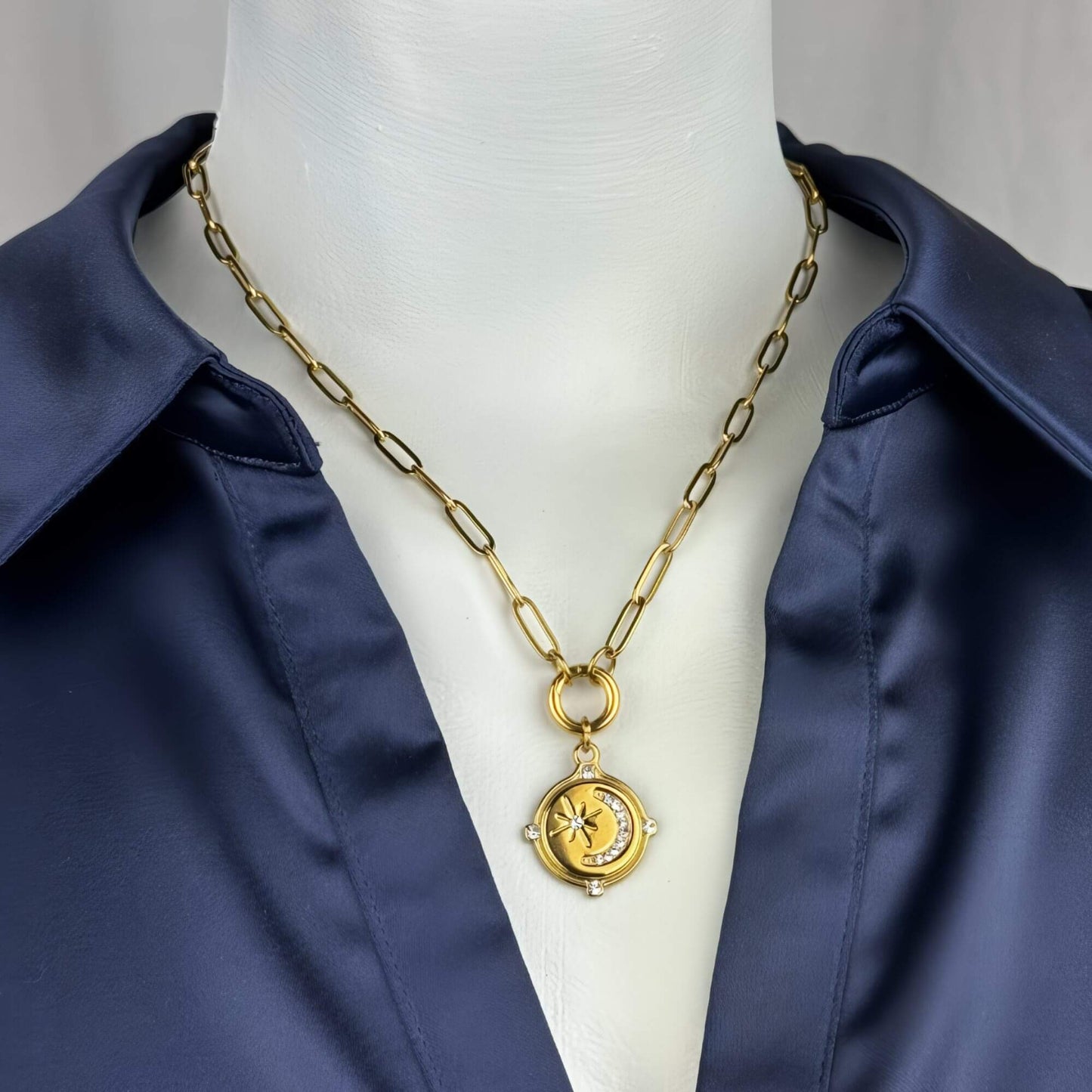 A gold-toned paperclip chain necklace with a circular pendant featuring a crescent moon and star design, displayed on a white mannequin torso wearing a navy satin shirt.