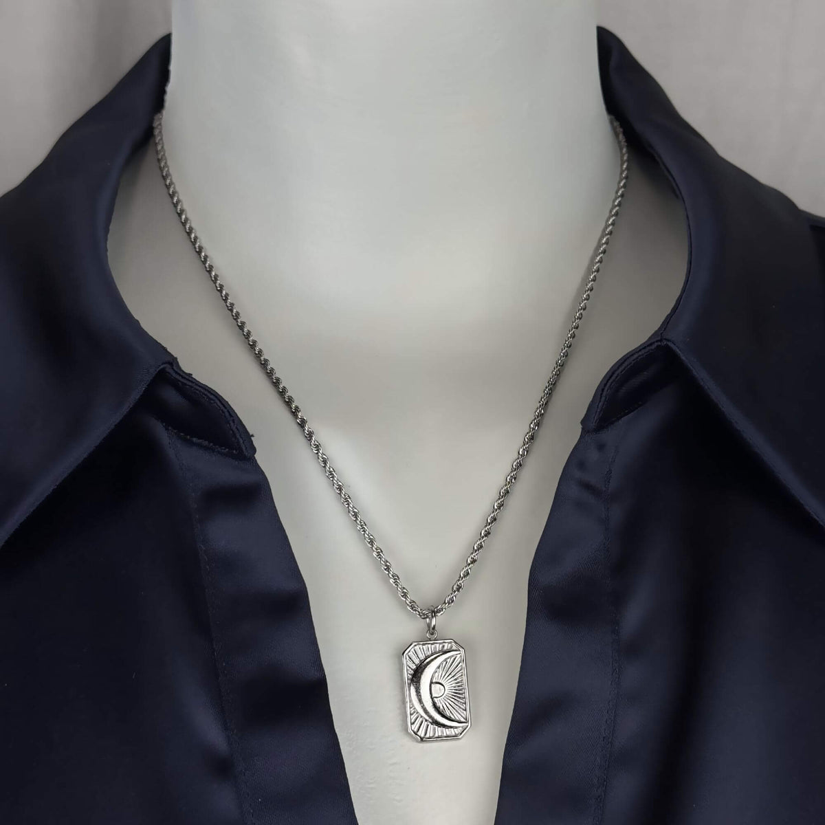 A silver-toned necklace with a rectangular pendant featuring a crescent moon and sun design, displayed on a white mannequin torso wearing a dark navy satin shirt.