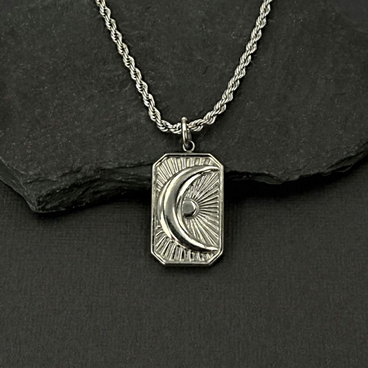 A silver-toned necklace with a rectangular pendant featuring a crescent moon and sunburst design, displayed on a dark textured surface with a black slate prop.