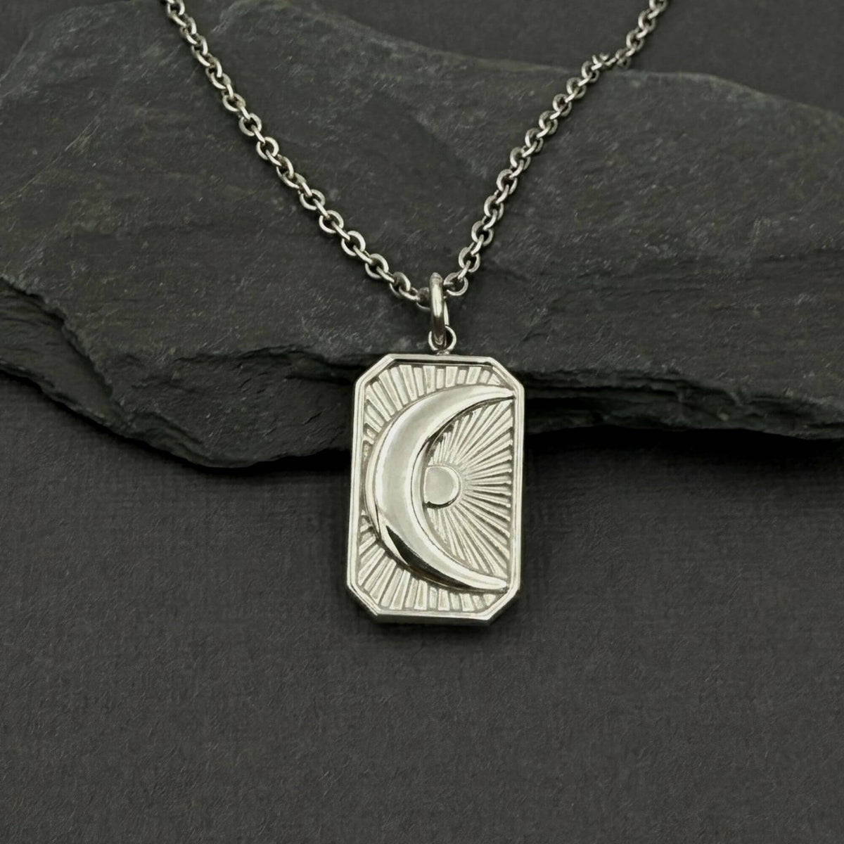 A silver-toned necklace with a rectangular pendant featuring an engraved crescent moon and sun design, displayed on a dark slate surface.
