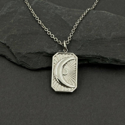 A silver-toned necklace with a rectangular pendant featuring an engraved crescent moon and sun design, displayed on a dark slate surface.