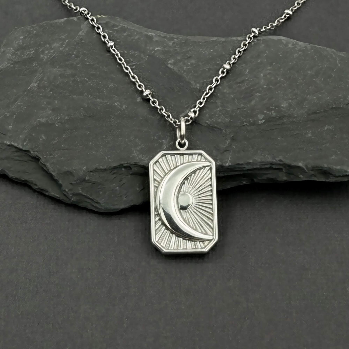 A silver pendant necklace featuring a crescent moon and sun design on a rectangular border, displayed on a dark slate rock against a dark gray background.