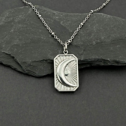 A silver pendant necklace featuring a crescent moon and sun design on a rectangular border, displayed on a dark slate rock against a dark gray background.