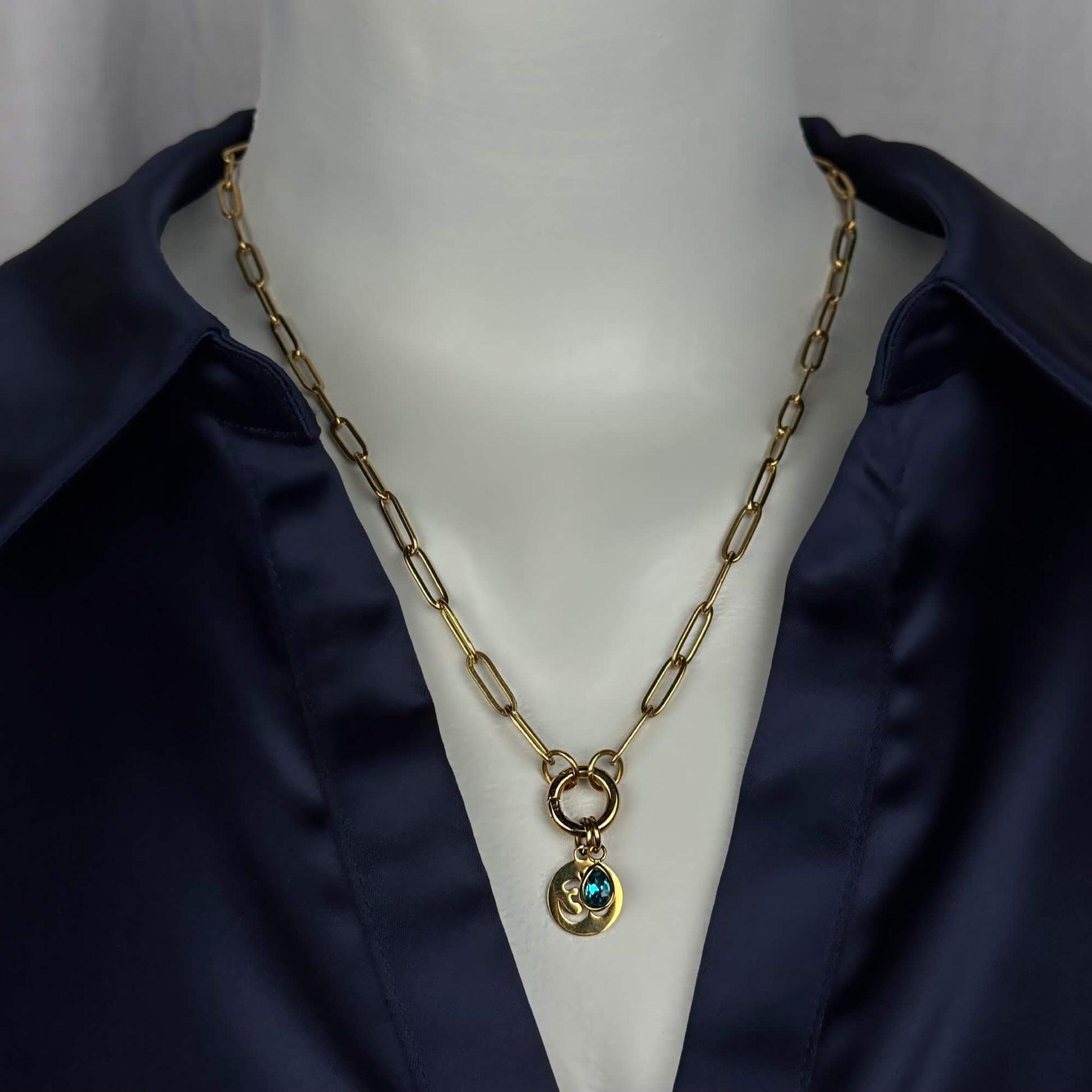 Gold paperclip chain necklace with a double-ring clasp and a small pendant featuring a blue-green stone, displayed on a white mannequin torso wearing a dark navy satin shirt.