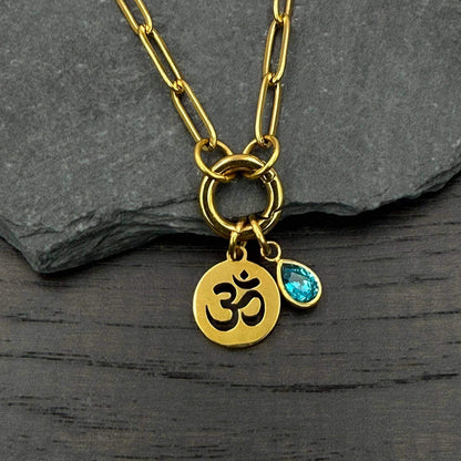 Gold-toned chain necklace with a round Om pendant and a small teardrop-shaped blue gemstone charm, displayed on a dark textured slate surface. The chain is a larger gold paperclip style.