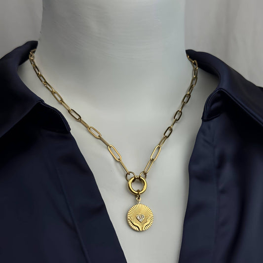 A gold paperclip chain necklace with a circular sunburst pendant featuring a small heart, displayed on a white mannequin torso wearing a dark navy satin shirt.