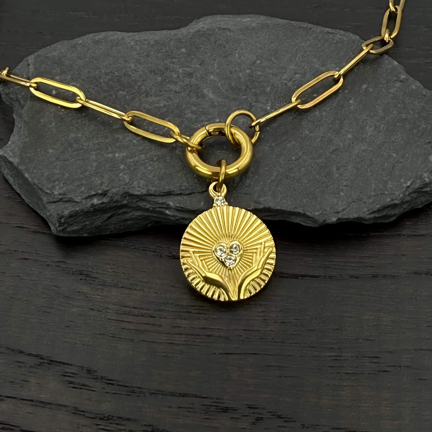 A gold pendant paperclip chain necklace with a circular sunburst charm featuring a small heart-shaped crystal and two hands Claddagh medallion, displayed on a dark textured stone and dark wood surface.